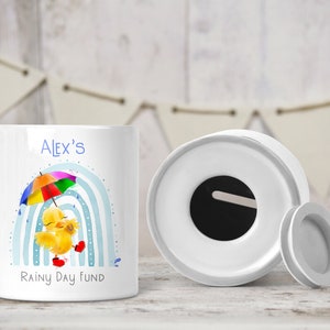 May include: White ceramic money box with a yellow duck holding an umbrella and a rainbow design. The money box has a slot on the top and says "Alex's Rainy Day Fund".
