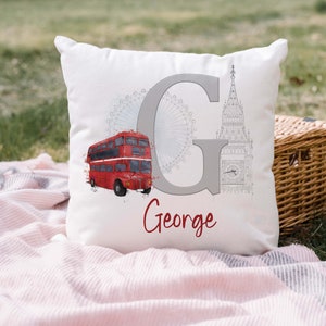 May include: A white square throw pillow featuring a large grey letter G, a red double-decker bus, a grey outline of the London Eye, and a grey sketch of Big Ben. The name George is printed in red cursive script below the letter G.
