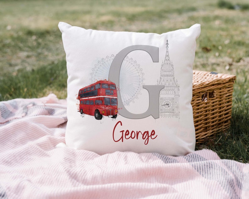 May include: A white square throw pillow featuring a large grey letter G, a red double-decker bus, a grey outline of the London Eye, and a grey sketch of Big Ben. The name George is printed in red cursive script below the letter G.