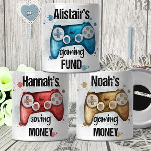 May include: Three white ceramic money boxes with colourful watercolour designs of video game controllers. The money boxes are labelled "Alistair's gaming FUND", "Hannah's saving MONEY", and "Noah's gaming MONEY".