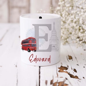 May include: A white ceramic money box featuring a large grey letter E, a red double-decker bus, a light blue London Eye illustration, and a grey Big Ben clock tower sketch. The name Edward is printed in red script font below the letter E.