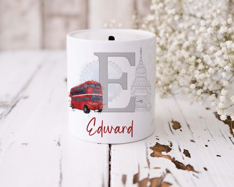 May include: A white ceramic money box featuring a large grey letter E, a red double-decker bus, a light blue London Eye illustration, and a grey Big Ben clock tower sketch. The name Edward is printed in red script font below the letter E.