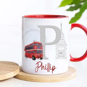 May include: A white ceramic mug with a red interior and handle, featuring a large grey letter P, a red double-decker bus, and a grey line drawing of the London Eye and Big Ben. The name Phillip is printed in red script at the bottom.