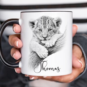 May include: White ceramic mug with a black handle. The mug features a black and white illustration of a lion cub on a branch. The name "Thomas" is written in cursive at the bottom of the mug.