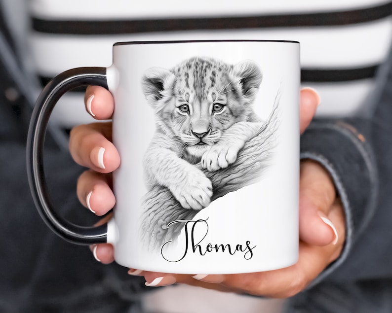 May include: White ceramic mug with a black handle. The mug features a black and white illustration of a lion cub on a branch. The name "Thomas" is written in cursive at the bottom of the mug.