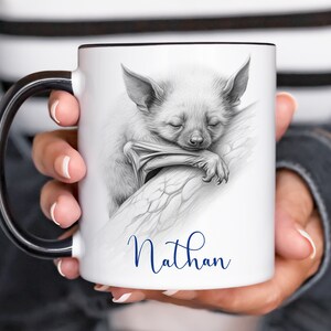May include: White ceramic mug with a black handle. The mug features a black and white pencil drawing of a bat sleeping on a branch. The name "Nathan" is written in blue cursive below the image.