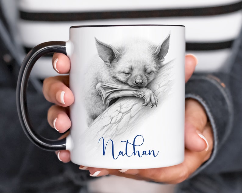 May include: White ceramic mug with a black handle. The mug features a black and white pencil drawing of a bat sleeping on a branch. The name "Nathan" is written in blue cursive below the image.