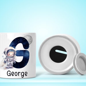 May include: White ceramic money box with a removable lid. The front of the box features a blue and black illustration of an astronaut and the letter G. The name George is printed below the illustration.