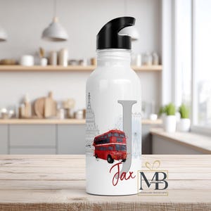 May include: A white water bottle with a black lid featuring a large grey letter J, a red double-decker bus illustration, and the name Jax written in red script. The design includes faint grey outlines of London landmarks like Big Ben.