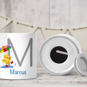 May include: White ceramic money box with a slot on the top. The box features a cartoon chick holding an umbrella and the letter "M" with the name "Marcus" printed below.