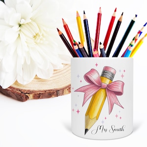 May include: A white pencil holder with a yellow pencil design and a pink bow, filled with colorful pencils. The pencil holder has the text "Mrs Smith" in black script. A white flower and a wooden slice are in the background.