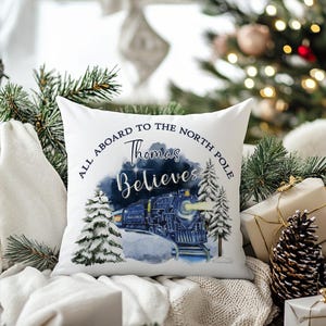 May include: White square pillow with a Christmas train design. The pillow features a blue train with the text "All Aboard to the North Pole, Thomas Believes." Snowy pine trees and a winter scene complete the design.