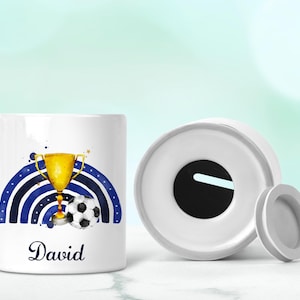 May include: White ceramic money box with a blue and white watercolour rainbow design, a gold trophy, and a football. The name "David" is written in blue below the design.