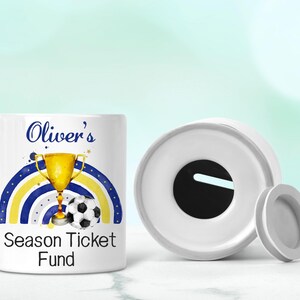 May include: White ceramic money box with a blue, yellow and white rainbow design, a gold trophy and a football. The money box has a slot on the top and says "Oliver's Season Ticket Fund".
