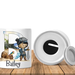 May include: White ceramic money box with a removable lid. The box features a colourful illustration of a pirate character with a sword, a parrot, and a treasure chest. The name "Bailey" is printed on the front of the box.