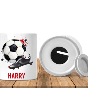 May include: White ceramic money box with a black and white football and a black football boot. The money box has a slot on the top and a lid. The money box is decorated with red and black paint splatters and black stars. The name "HARRY" is written in red letters on the front of the money box.