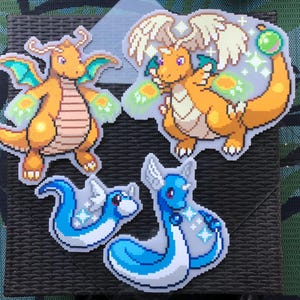 May include: Four colorful pixel art stickers featuring dragon-like creatures. Two are orange with wings, one with a green orb. The other two are blue, one curled up and the other with a long body. The stickers are on a textured surface.
