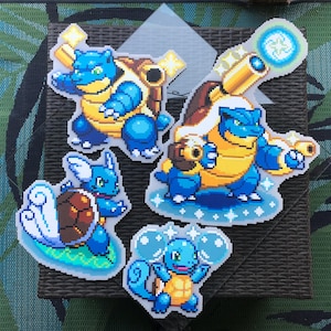 May include: Four pixel art stickers depicting blue and yellow turtle-like creatures. The stickers, with white borders, are arranged on a dark brown surface. The creatures are in different poses, some with water effects, reminiscent of a popular franchise.