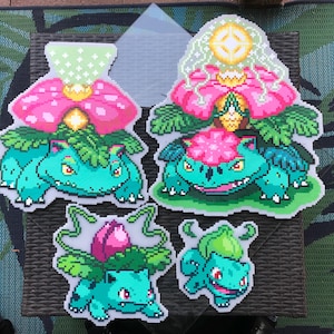 Könnte beinhalten: Vier Pixel-Art-Figuren der Pokémon-Figur Bisasam. Die Figuren sind in verschiedenen Größen und zeigen Bisasam in verschiedenen Entwicklungsstadien, mit grünen Körpern, rosa Blüten und grünen Blättern. Die Figuren befinden sich auf einer dunkelbraunen Oberfläche.