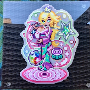 May include: Pixelated sticker of a cartoon princess with blonde hair, a crown, and a star-shaped wand. The princess wears a purple dress and holds a paintbrush with a rainbow palette. The sticker has a pink and green colour scheme.
