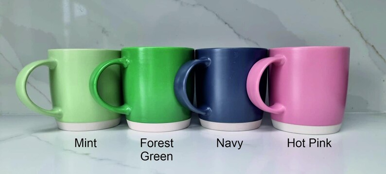 K&ouml;nnte beinhalten: Vier Keramikbecher in verschiedenen Farben: Mintgr&uuml;n, Waldgr&uuml;n, Marineblau und Pink. Jeder Becher hat einen wei&szlig;en Streifen am Boden.