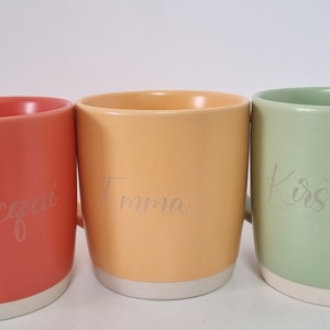 K&ouml;nnte beinhalten: Drei Keramikbecher mit wei&szlig;en R&auml;ndern und Griffen. Die Becher sind orange, gelb und gr&uuml;n. Die Namen "Jacqui", "Twanna" und "Kirsty" sind in die Becher graviert.