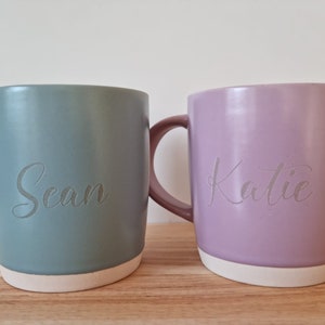 K&ouml;nnte beinhalten: Zwei Keramikbecher mit personalisierten Namen. Der Becher links ist in einem ged&auml;mpften Gr&uuml;nton gehalten und tr&auml;gt den Namen "Sean". Der Becher rechts ist in einem hellen Lila gehalten und tr&auml;gt den Namen "Katie".