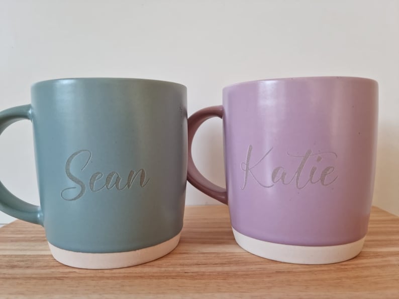 K&ouml;nnte beinhalten: Zwei Keramikbecher mit personalisierten Namen. Der Becher links ist in einem ged&auml;mpften Gr&uuml;nton gehalten und tr&auml;gt den Namen "Sean". Der Becher rechts ist in einem hellen Lila gehalten und tr&auml;gt den Namen "Katie".