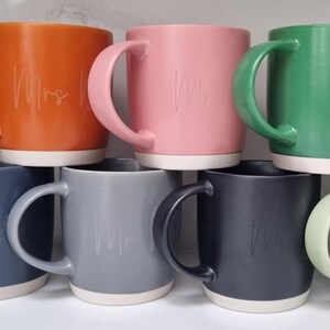 K&ouml;nnte beinhalten: Set aus 7 Keramikbechern in verschiedenen Farben, jede mit einem wei&szlig;en Rand und einem wei&szlig;en Boden. Die Becher haben das Wort "Frau" in kursivschrift darauf geschrieben.