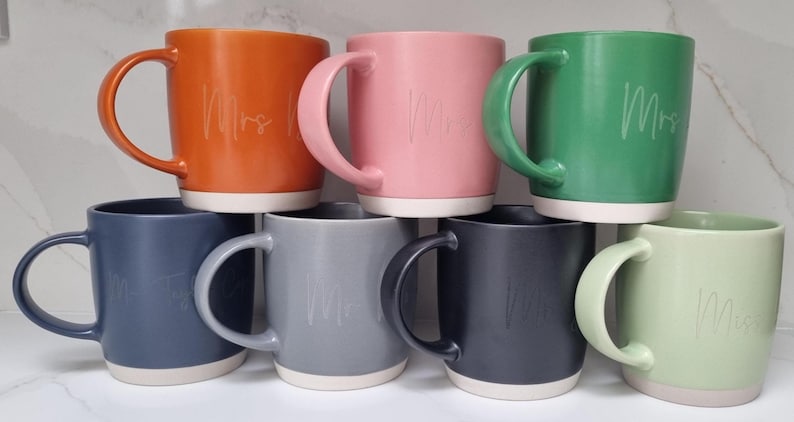 K&ouml;nnte beinhalten: Set aus 7 Keramikbechern in verschiedenen Farben, jede mit einem wei&szlig;en Rand und einem wei&szlig;en Boden. Die Becher haben das Wort "Frau" in kursivschrift darauf geschrieben.