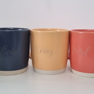 K&ouml;nnte beinhalten: F&uuml;nf Keramikbecher mit verschiedenen Glasuren und personalisierten Namen: Louis, Olivia, Kerry, Noel und NPO. Die Becher haben einen wei&szlig;en Rand und Boden.