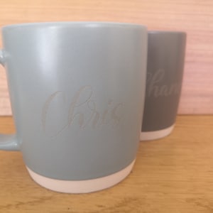 K&ouml;nnte beinhalten: Zwei Keramikbecher mit wei&szlig;en R&auml;ndern. Der Becher links ist hellblau und hat den Namen "Chris" eingraviert. Der Becher rechts ist grau und hat den Namen "Sharon" eingraviert.