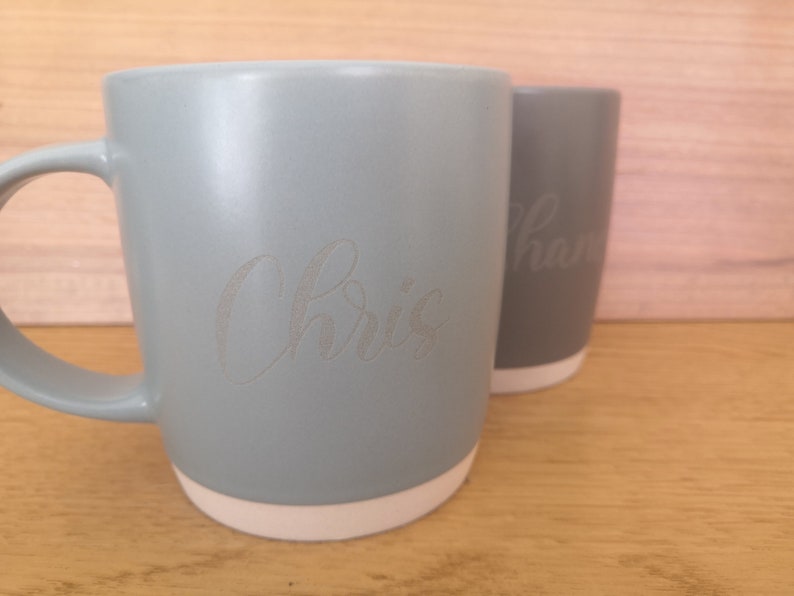 K&ouml;nnte beinhalten: Zwei Keramikbecher mit wei&szlig;en R&auml;ndern. Der Becher links ist hellblau und hat den Namen "Chris" eingraviert. Der Becher rechts ist grau und hat den Namen "Sharon" eingraviert.