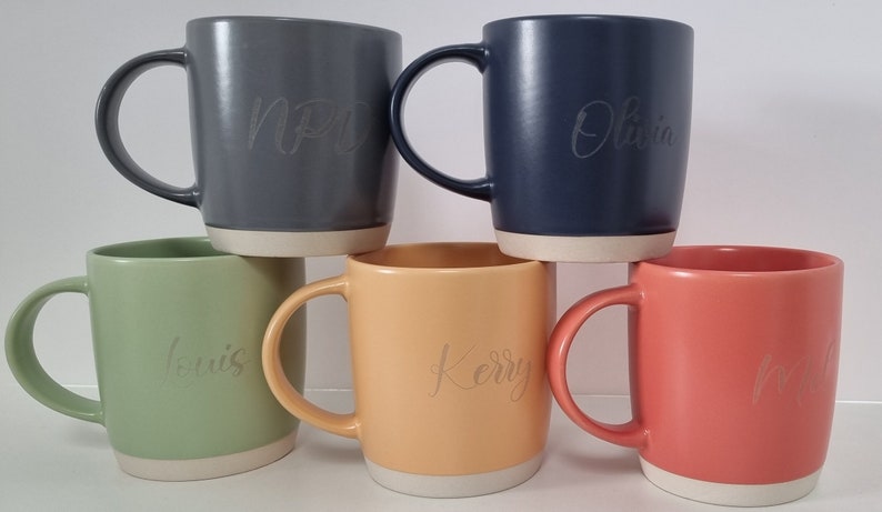K&ouml;nnte beinhalten: F&uuml;nf Keramikbecher mit verschiedenen Glasuren. Die Becher sind pyramidenf&ouml;rmig gestapelt. Die Becher sind mit den Namen "Olivia", "Kerry", "Luis", "NP" und "Lori" personalisiert.