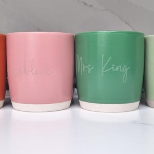 K&ouml;nnte beinhalten: Vier Keramikbecher mit verschiedenen Glasuren und wei&szlig;er Beschriftung. Die Becher sind mit "Ballentine", "Caleb", "Frau King" und "Frau Peters" beschriftet.