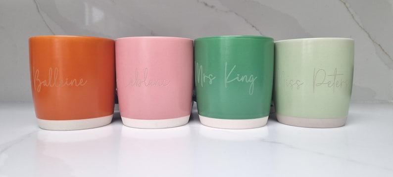 K&ouml;nnte beinhalten: Vier Keramikbecher mit verschiedenen Glasuren und wei&szlig;er Beschriftung. Die Becher sind mit "Ballentine", "Caleb", "Frau King" und "Frau Peters" beschriftet.