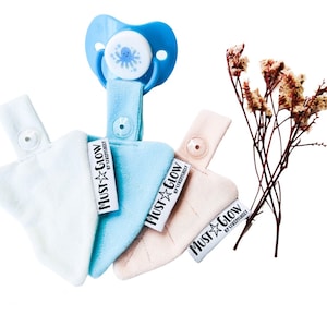 Op de afbeelding: Drie zachte, pluche, driehoekige bijtringen in blauw, roze en wit met een blauwe en witte octopus-speenclip. Elke bijtring heeft een label met de tekst "Must Glow by Siegfried & Roy".