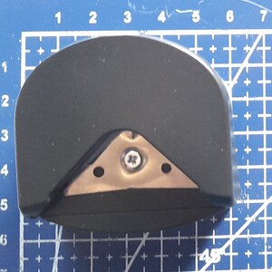 May include: A black and silver corner rounder tool with a triangular blade and a screw in the center. The tool is designed to round the corners of paper or cardstock.