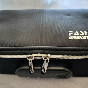 May include: Black rectangular case with a silver zipper and combination lock. The text "FASHION BASKETBALL" is printed in white on the top of the case. The case is designed to store items.