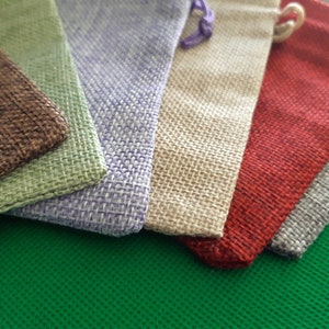 May include: Six small burlap pouches in various colors, including brown, green, purple, beige, red, and gray. The pouches are arranged in a stack, with the brown pouch on the bottom and the gray pouch on the top.