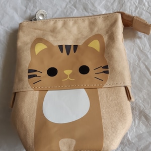 May include: A beige canvas pencil case with a cartoon cat design. The cat has brown fur, yellow ears, black eyes, and a white belly.