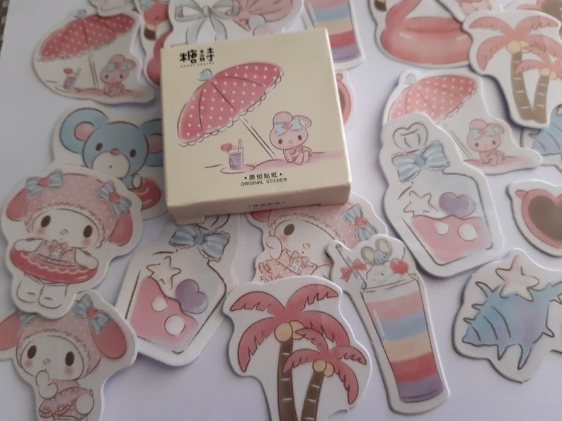 K&ouml;nnte beinhalten: Ein Set mit pink- und blaut&ouml;nigen Aufklebern, die ein Comic-Figur mit einer rosa Schleife und einem rosa Kleid zeigen. Die Aufkleber sind in verschiedenen Formen und Gr&ouml;&szlig;en, darunter Palmen, Muscheln und ein Parf&uuml;mflakon. Die Aufkleber sind in einer kleinen wei&szlig;en Schachtel verpackt, auf der "Original Sticker" steht.