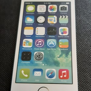 Puede incluir: Un iPhone blanco con una pantalla negra que muestra una variedad de aplicaciones, incluyendo Calendario, Fotos, Cámara, Clima, Reloj, Mapas, Vídeos, Notas, Recordatorios, Acciones, Centro de juegos, Quiosco, iTunes Store, App Store, Passbook, Brújula, Ajustes, Teléfono, Correo, Safari y Música.