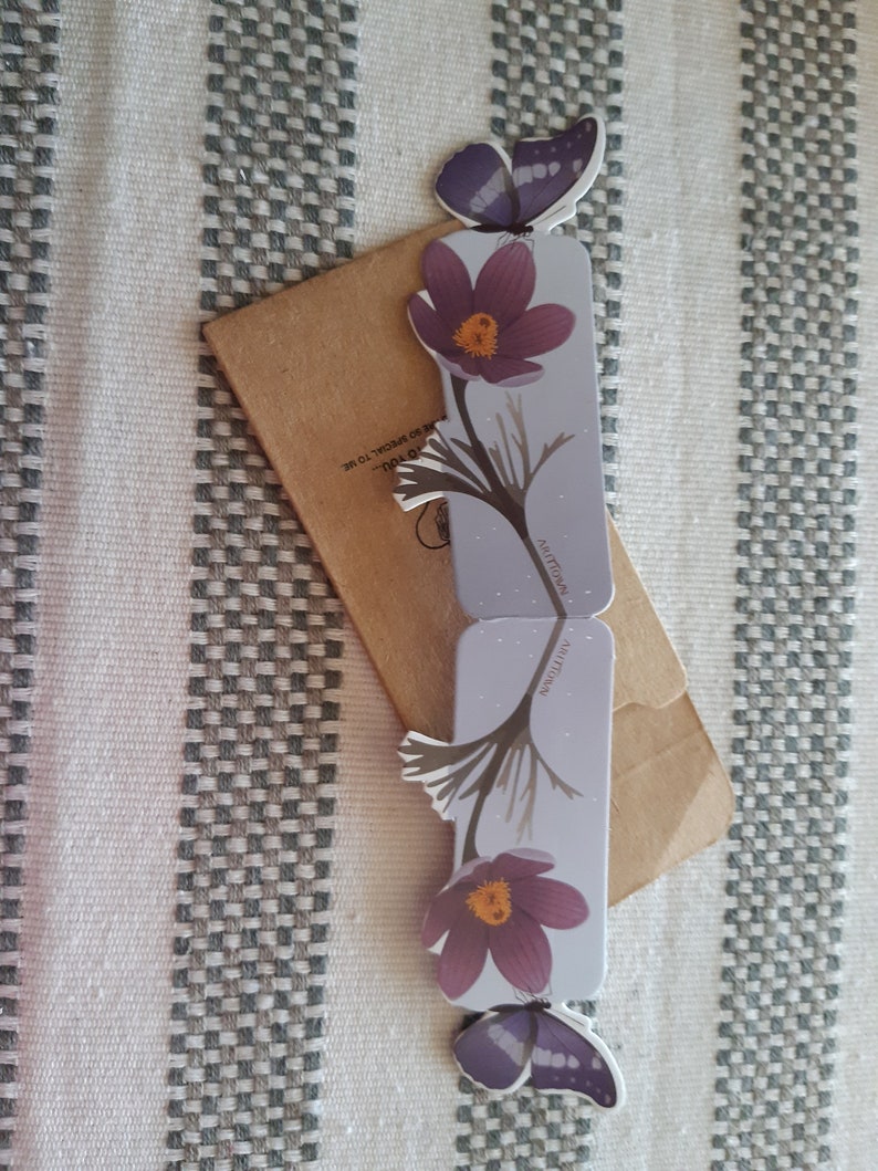 May include: Two purple and white butterfly bookmarks with a flower design. The bookmarks are on a brown envelope with the text "HOLO" and "281.700.0038" printed on it.