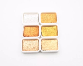 Puede incluir: Seis pastillas de pintura de acuarela en una bandeja de plástico blanco. Las pastillas contienen colores metálicos dorados, plateados y cobrizos.