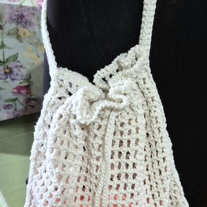 May include: A cream-colored, crocheted drawstring bag with a shoulder strap. The bag has a mesh-like pattern, and the interior is lined with a peach-colored fabric. The bag is hanging on a black surface.