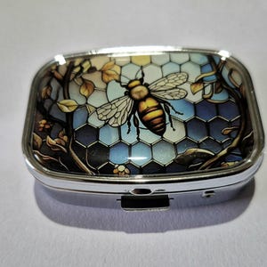 Może przedstawiać: Prostokątne, srebrne pudełko na pigułki z witrażowym wzorem przedstawiającym pszczołę. Pszczoła jest żółta i brązowa, na tle niebiesko-białego wzoru plastra miodu. Pudełko ma klapkę na zawiasach i bezpieczne zapięcie.