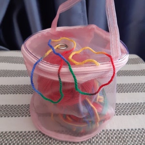 May include: Pink mesh yarn storage bag with a zipper closure and a handle. The bag contains colorful yarn balls.