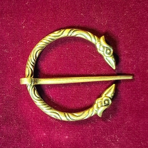May include: A gold-toned metal brooch with a circular design and two bird-shaped clasps. The brooch has a textured surface with a wavy pattern.