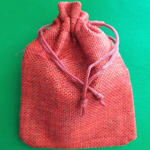 May include: A red drawstring bag made of burlap fabric. The bag is closed with a knotted cord.